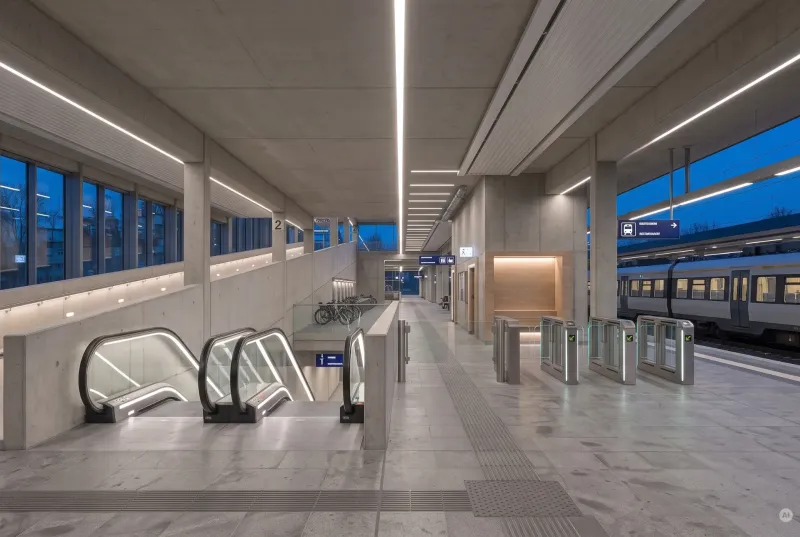Barrierefreier Bahnhofsterminal aus Sichtbeton mit hellen LED-Lichtbändern, Rolltreppen, Ticket-Zugangsschranken und einem einfahrenden Zug am Bahnsteig.