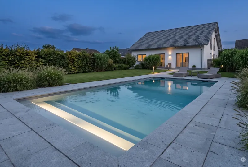 Rechteckiger Swimmingpool im Garten eines Einfamilienhauses bei Dämmerung mit leuchtender LED-Stufenbeleuchtung im Wasser und Natursteinumrandung.