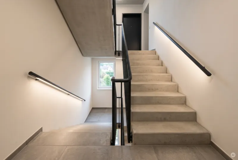 Minimalistische Sichtbetontreppe mit einem schwarzen wandmontierten LED-Lichtprofil als seitliche Stufen- und Orientierungsbeleuchtung.