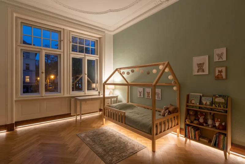 Gemütliches Kinderzimmer in einem Altbau mit hohen Decken, Fischgrätparkett, Hausbett und dezentem, warmweißem LED-Orientierungslicht entlang der Sockelleiste am Abend.