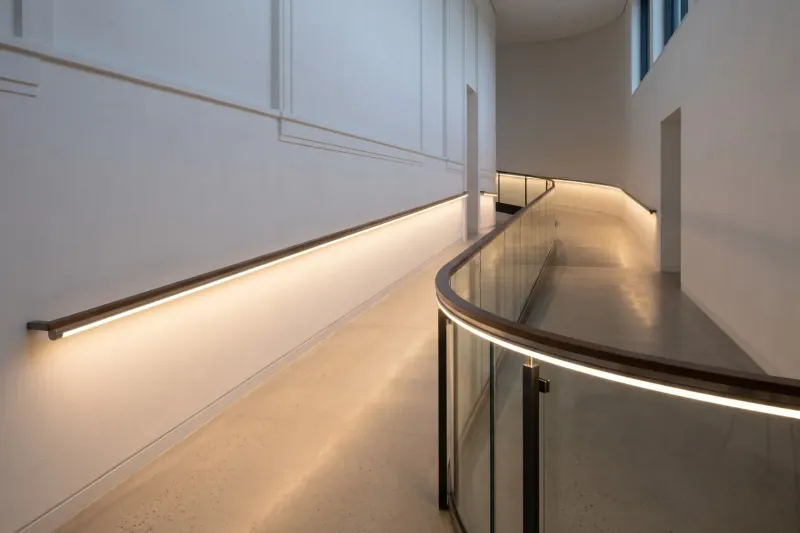 Ein moderner, heller Flur mit einer sanft ansteigenden Rampe und einer durchgehenden, warmweißen LED-Lichtlinie unter dem Handlauf an der weißen Wand und dem Glasgeländer.